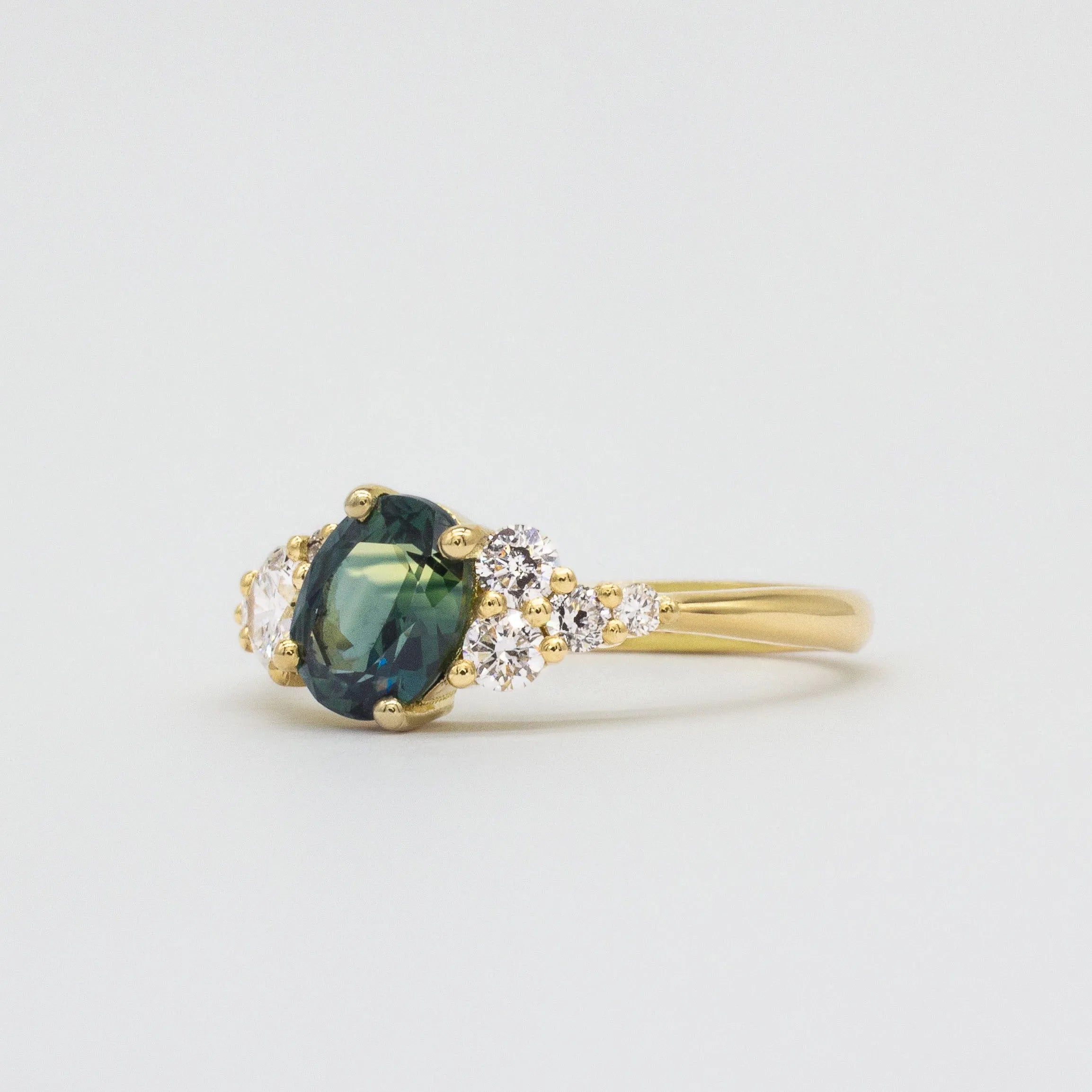 Gold ring with a blue green sapphire and diamonds on a light gray background