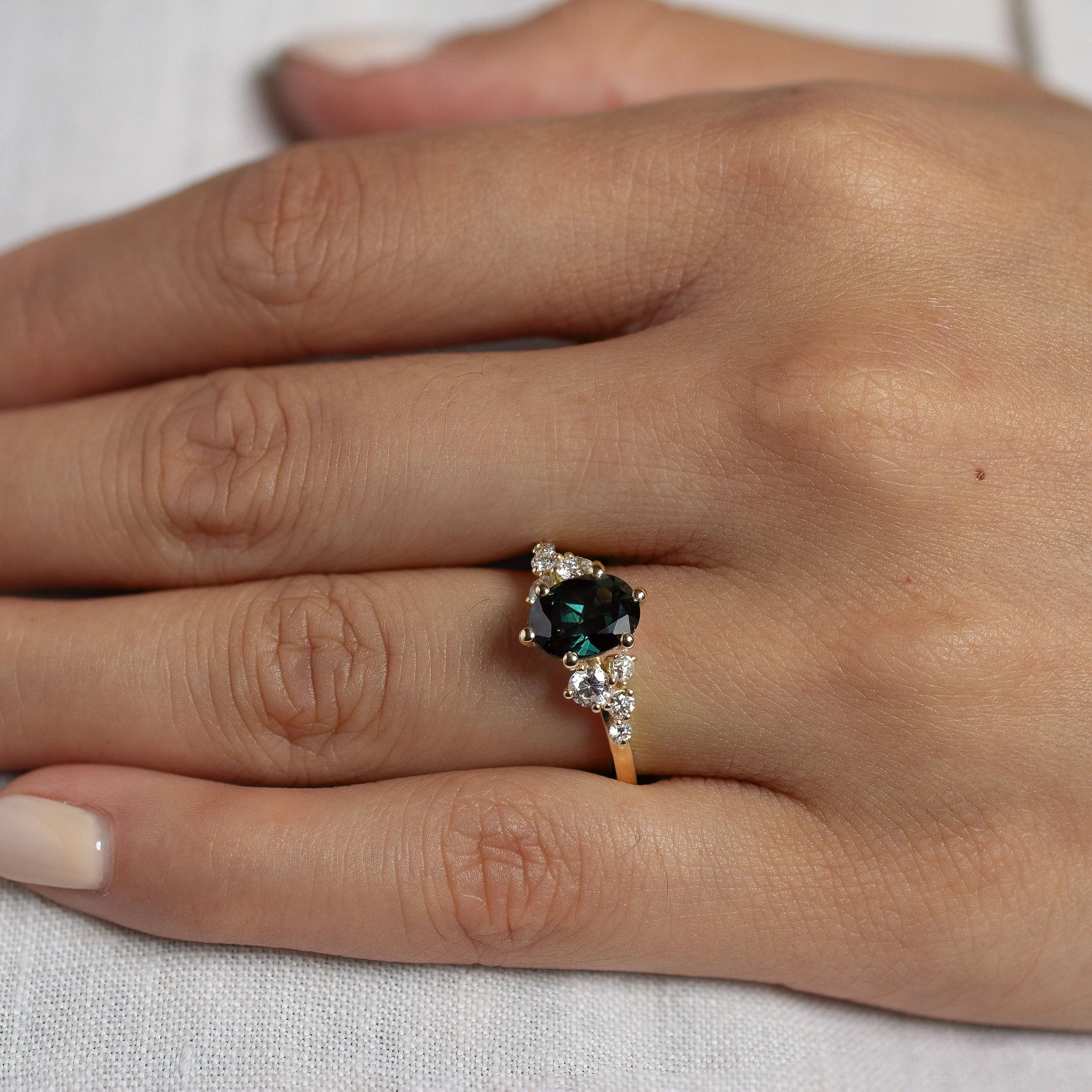 close up of hand wearing 6 prong blue green sapphire engagement ring with diamond cluster