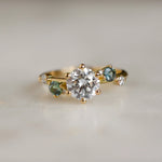 gold engagement ring with round diamond and blue and green sapphires with blurred background