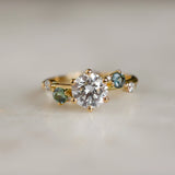 gold engagement ring with round diamond and blue and green sapphires with blurred background