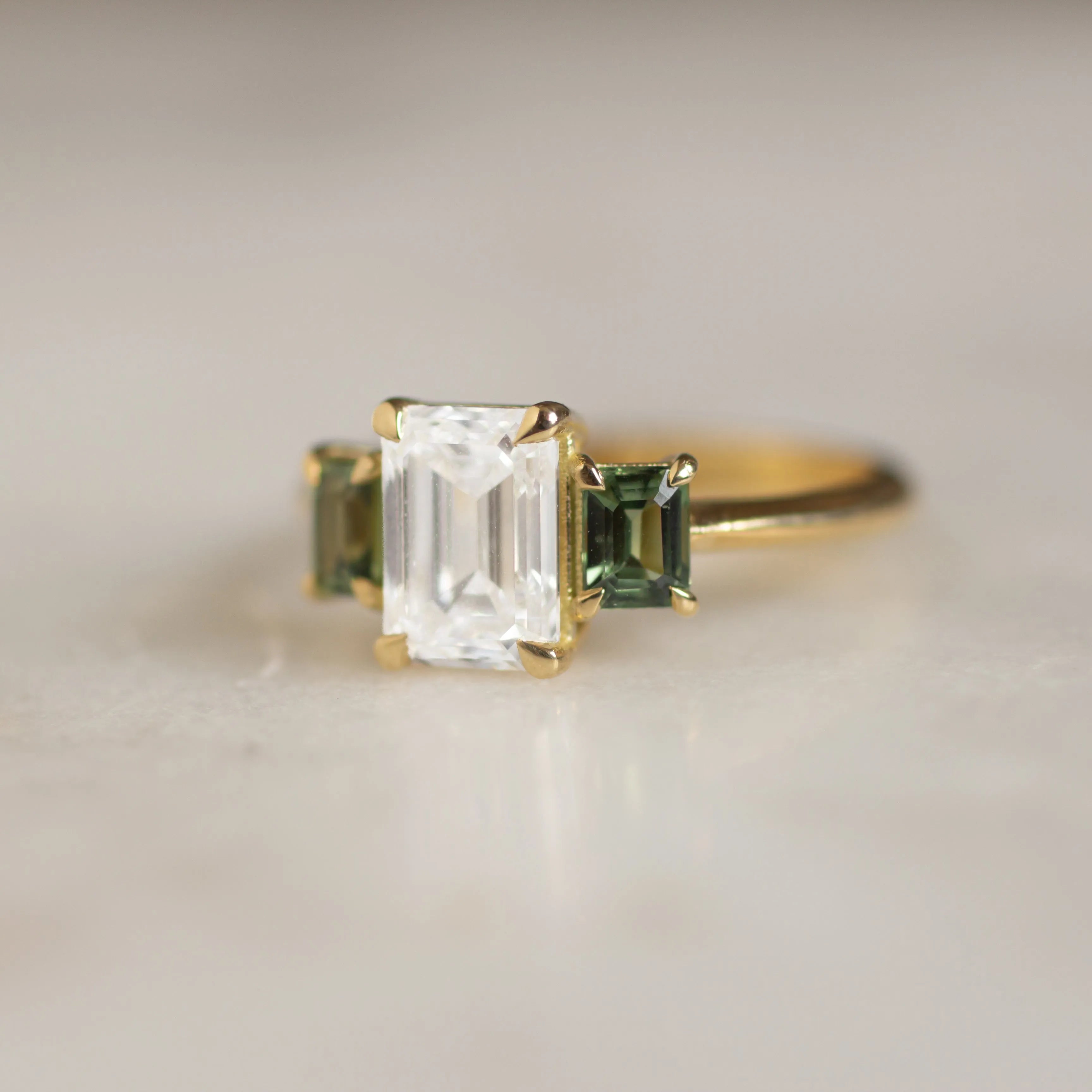 engagement ring with rectangle diamond and green sapphires with gold band and claws with blur background