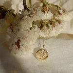Gold compass necklace with a round pendant on a white fabric background with cherry blossoms.