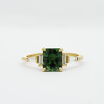 Gold engagement ring with a green cushion cut sapphire and tapered baguetter diamonds on a white background