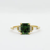 Gold engagement ring with a green cushion cut sapphire and tapered baguetter diamonds on a white background