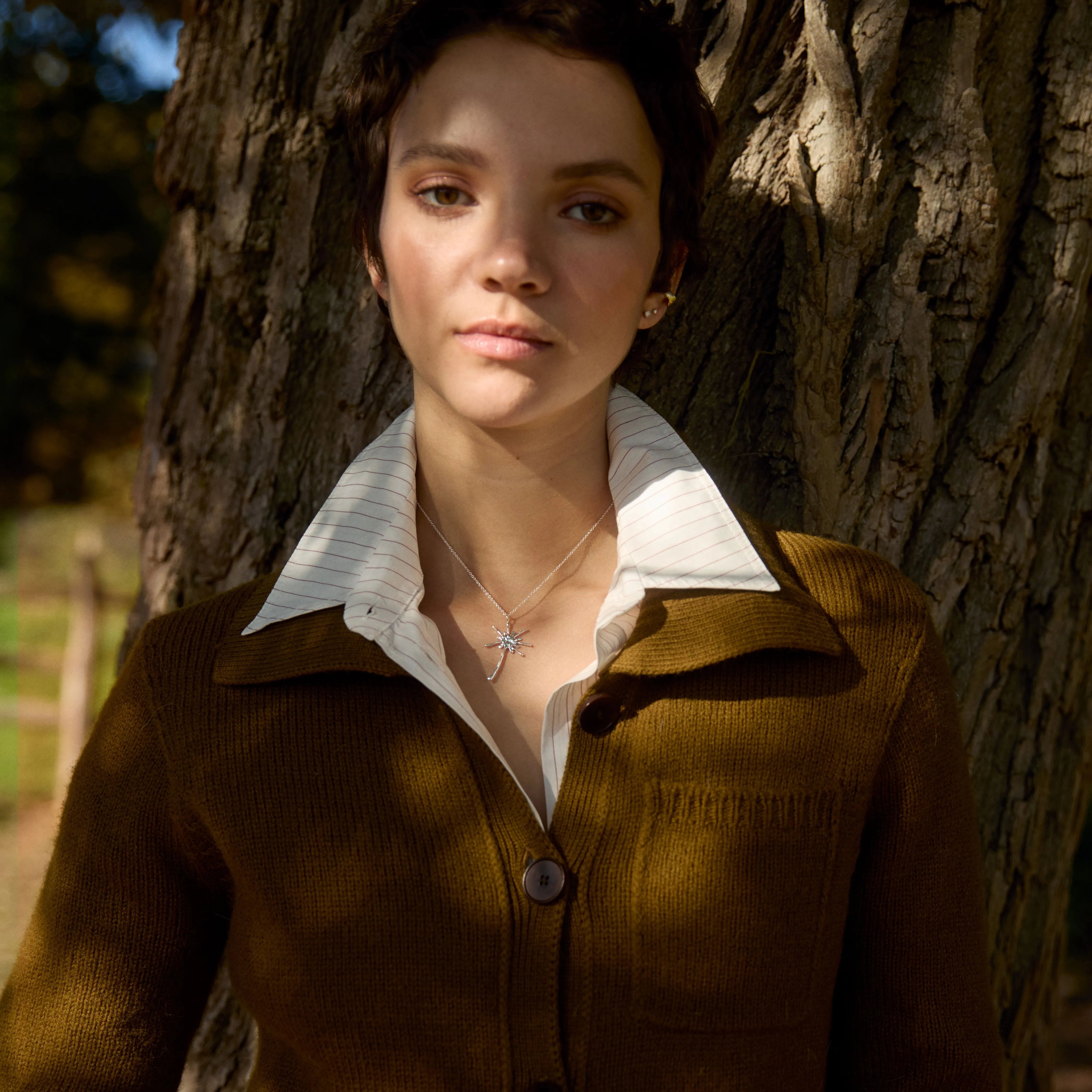 Woman standing next to a tree wearing diamond studs, dandelion necklace, brown cardigan and white shirt.