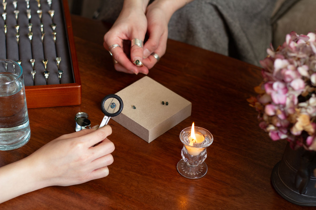 hand holding magnifying tool holding a blue sapphire, with another set of hands holding an engagement ring