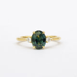 Gold ring with a teal green sapphire and smaller diamonds on a white background