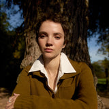 Woman in a brown sweater standing in front of a tree wearing a number necklace
