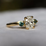 gold engagement ring with large oval diamond and small round green sapphires with blurred background