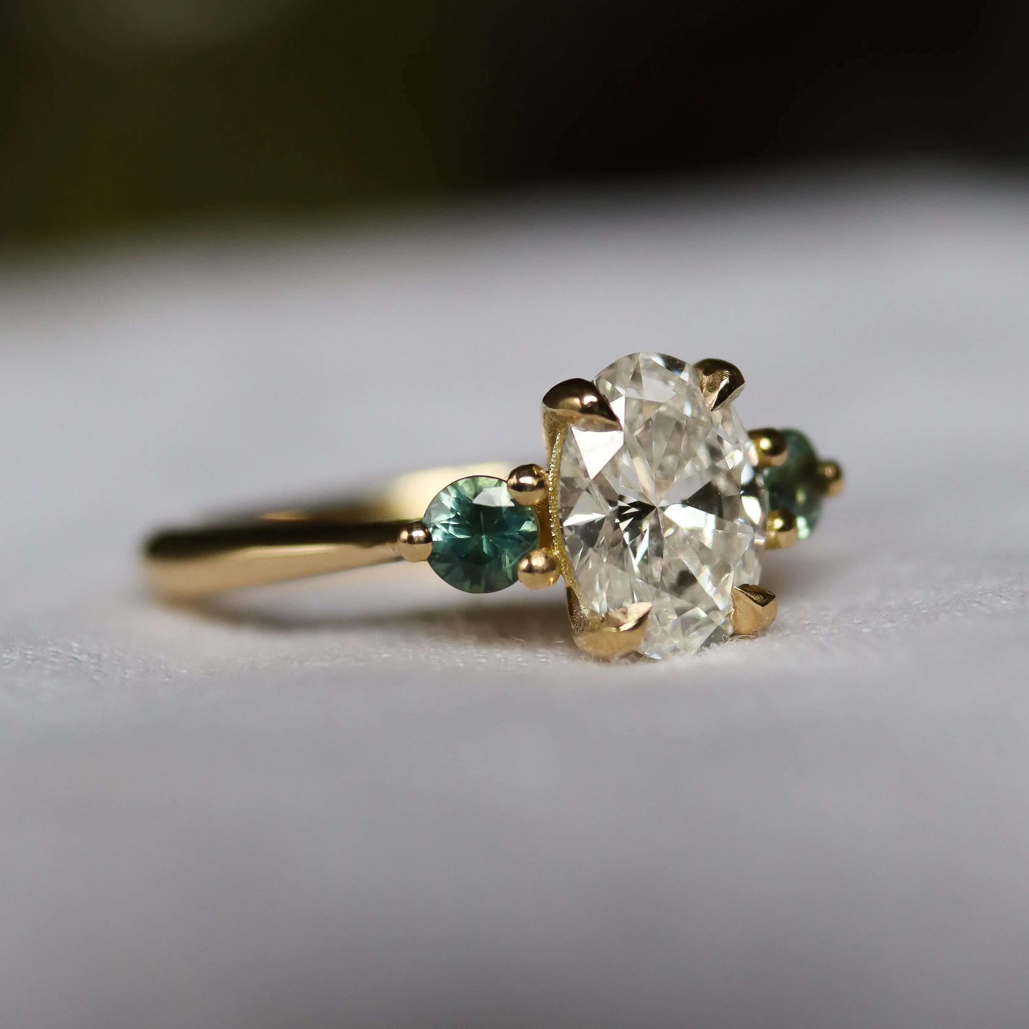 gold engagement ring with large oval diamond and small round green sapphires with blurred background