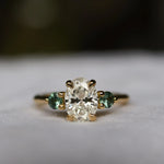 large diamond oval engagement ring with green sapphires and blurred background