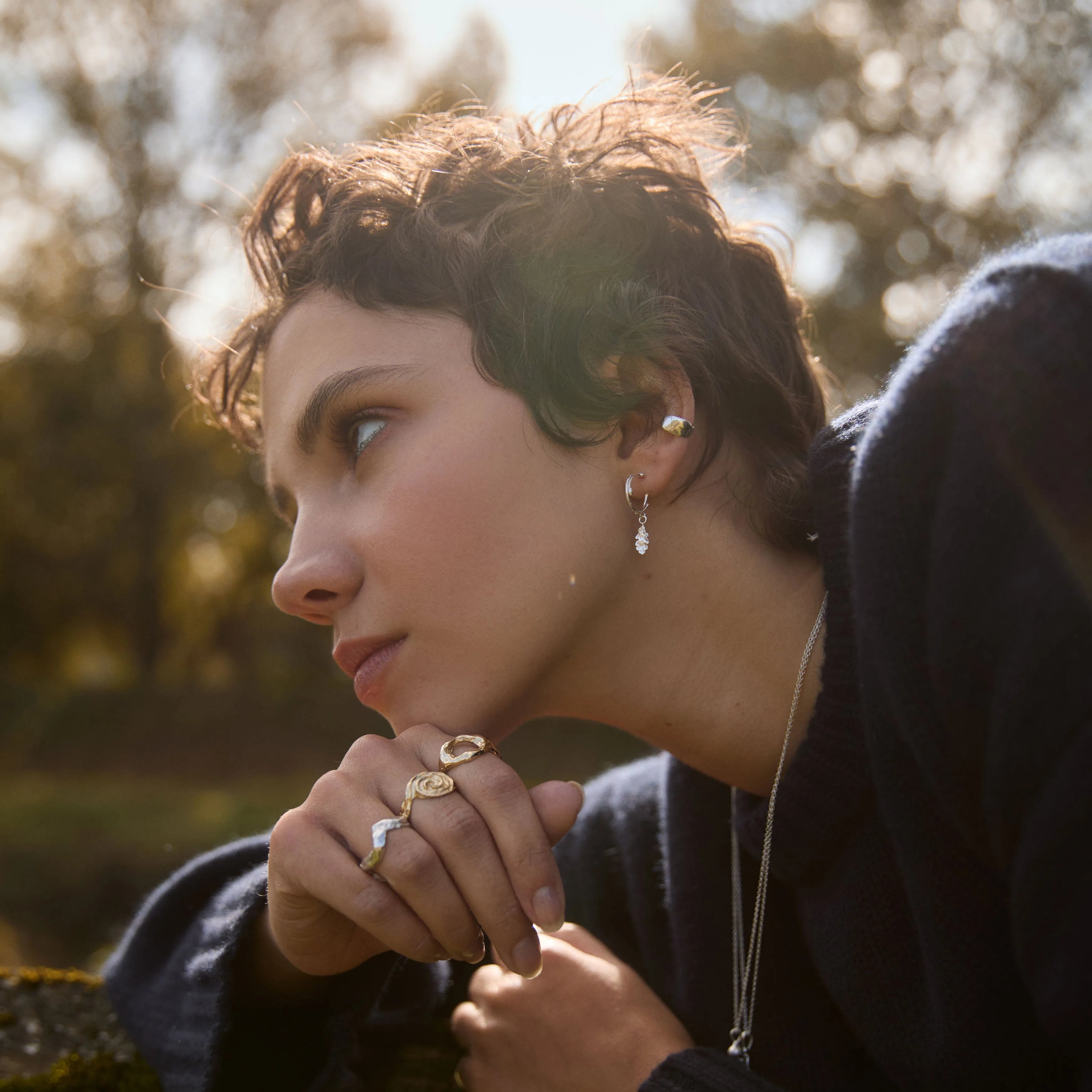 Woman with short hair and earrings, wearing a dark sweater, sitting outdoors with trees in the background.
