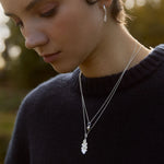 Close-up of a woman wearing a silver acorn and oak leaf necklaces and earrings with a blurred natural background