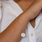 Silver bracelet with a round charm on a wrist, worn by a person in a white shirt.