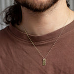 close up of gold raw rectangle pendant necklace worn by bearded man wearing brown tshirt