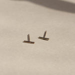 solid yellow gold bar stud earrings on beige surface with leaf shadows