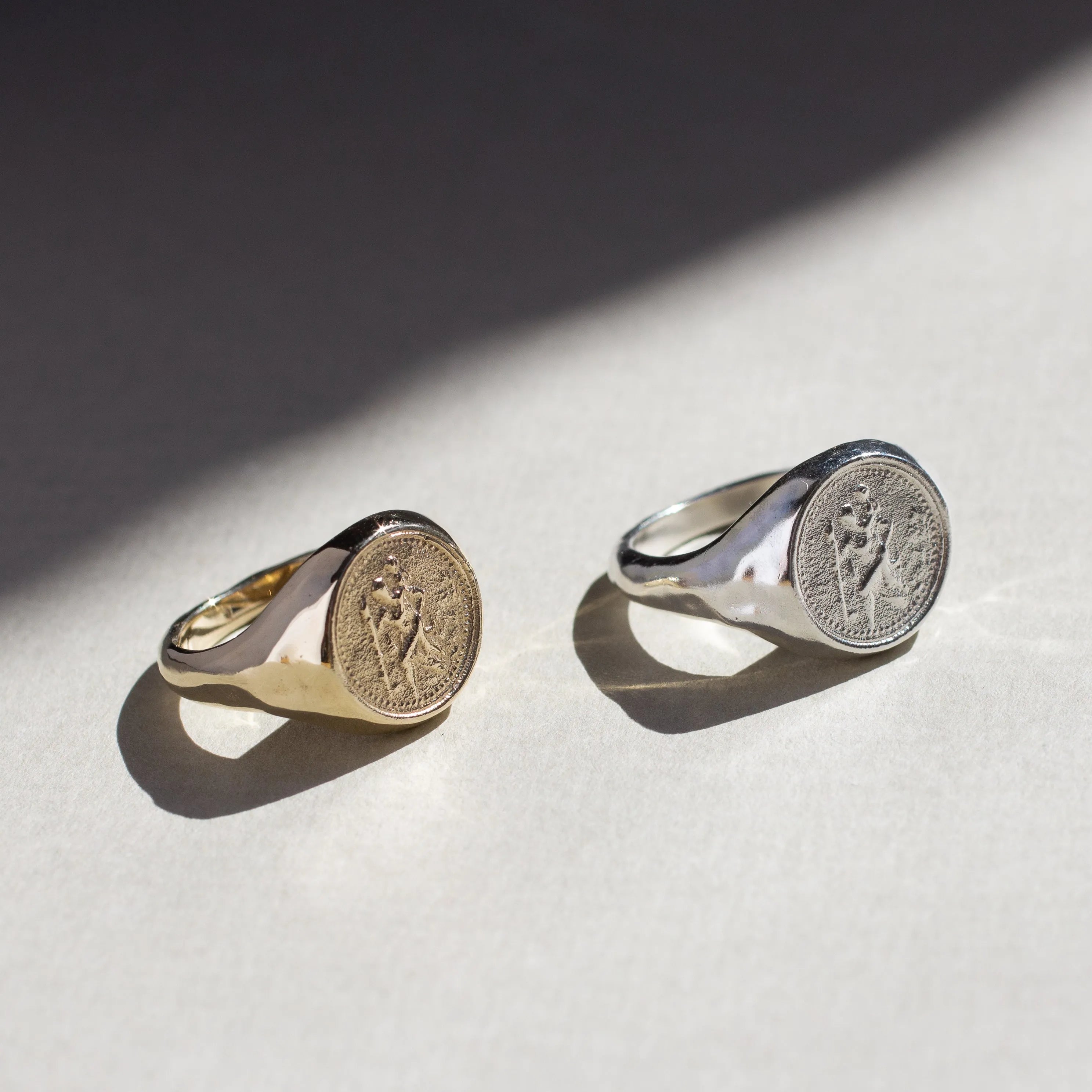 Two st christopher rings, one st christopher ring in gold and one silver, on a light surface with a soft shadow.