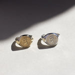 Two saint christopher signet rings, one gold and one silver, on a light surface with a soft shadow.