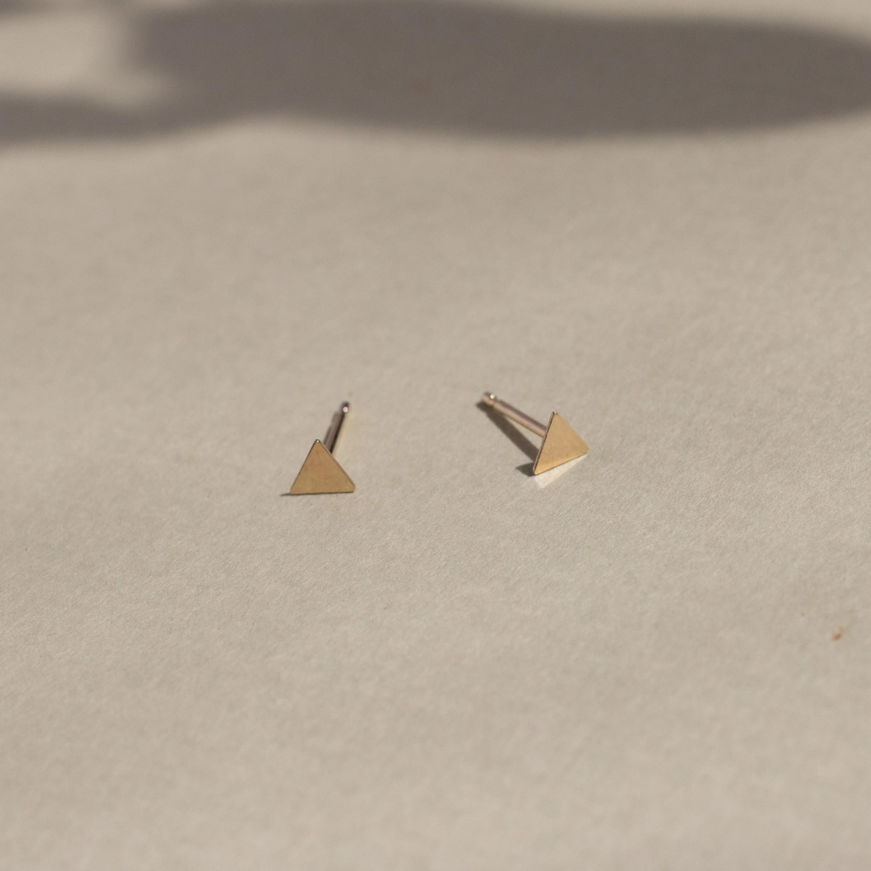 solid yellow gold triangle stud earrings on beige surface with leaf shadows
