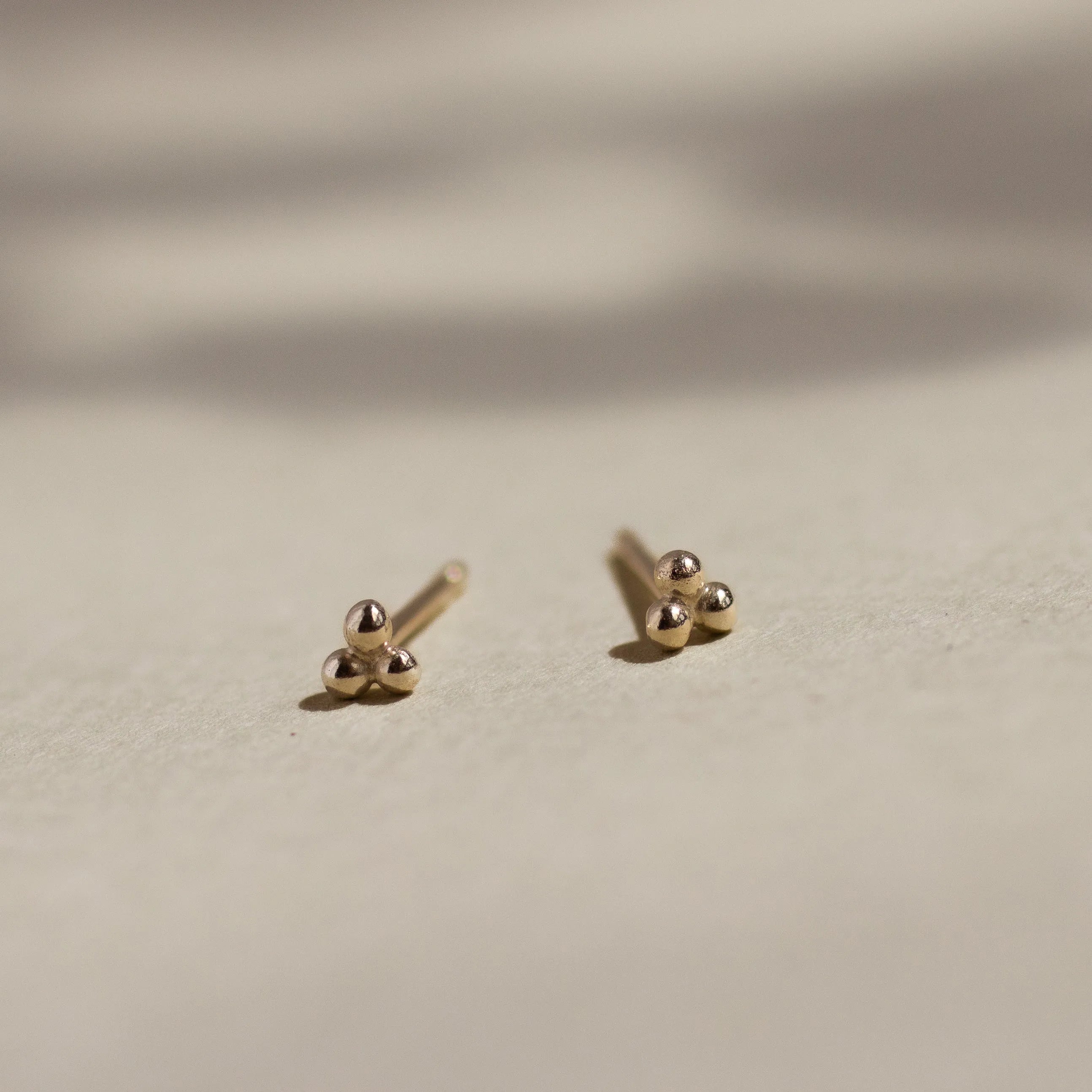 yellow gold triple dot stud earrings on beige surface with leaf shadows