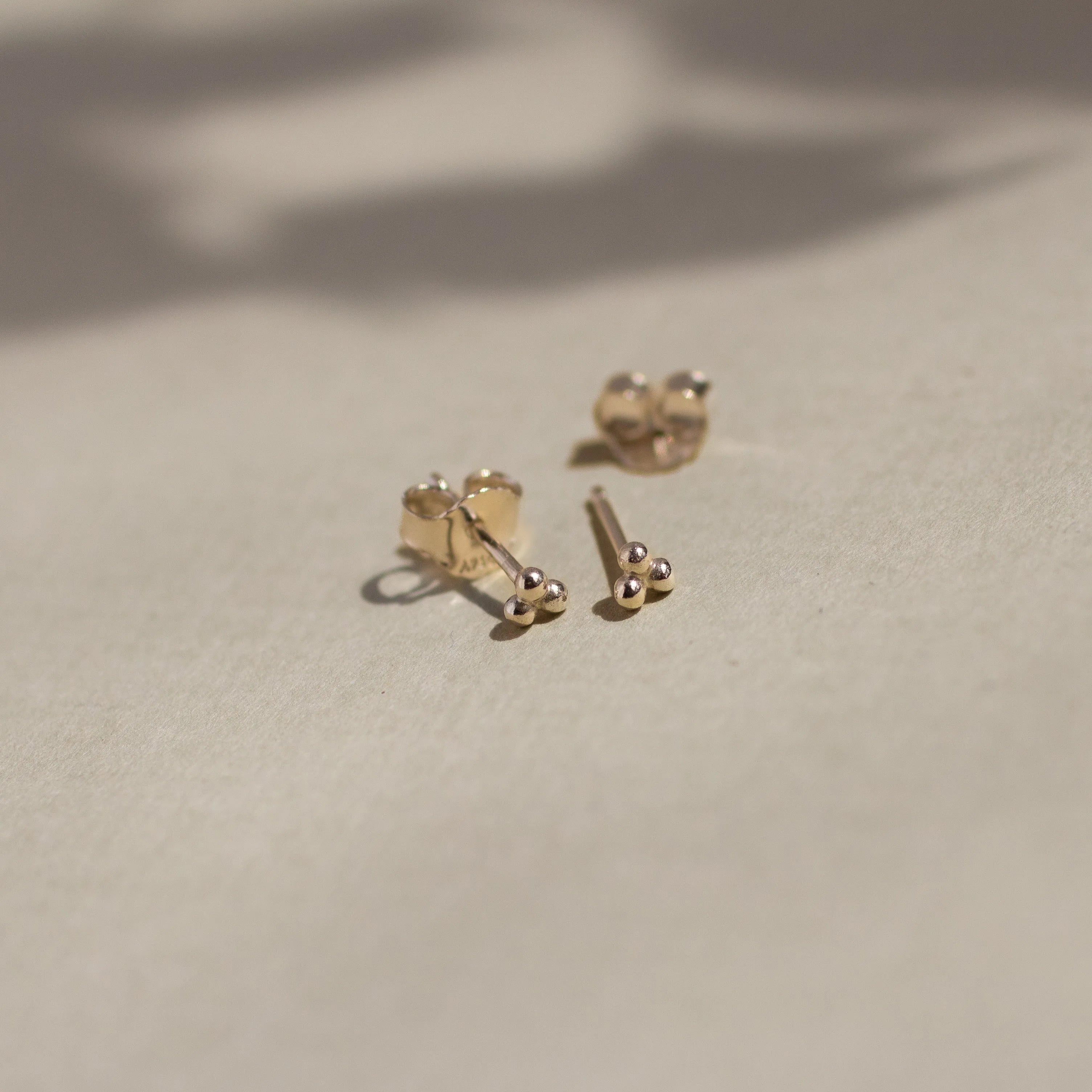 solid yellow gold triple dot stud earrings with butterfly backs on beige surface with leaf shadows