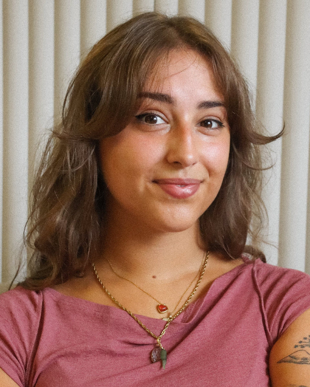 Woman with wavy hair wearing a pink top and multiple necklaces against a neutral background
