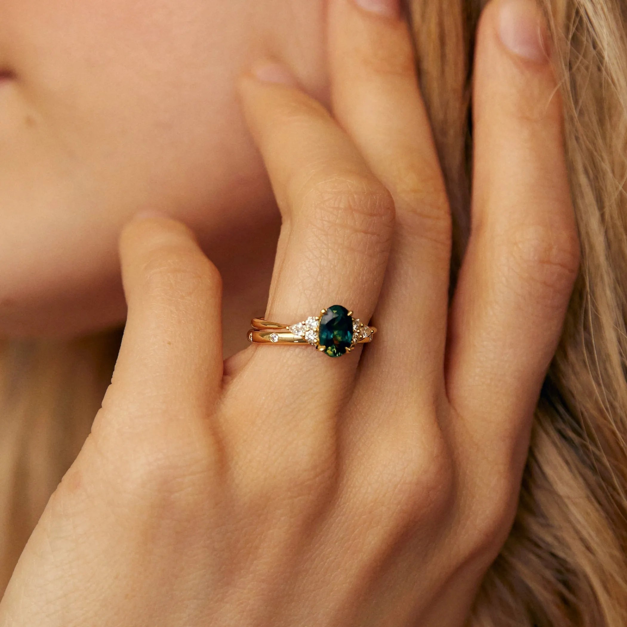 Close-up of a hand wearing a gold ring with a green gemstone, touching hair.