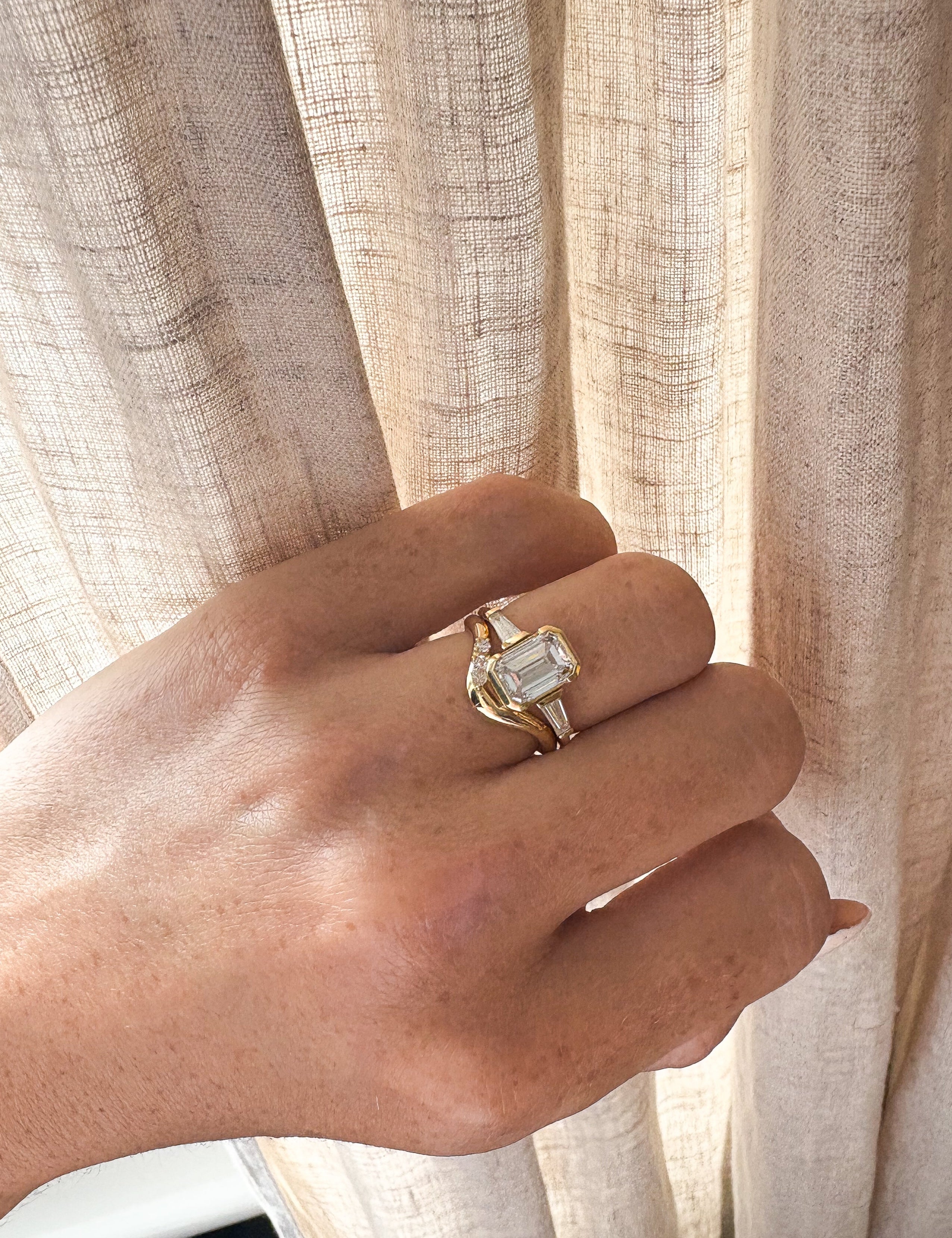 Hand wearing a gold ring with a diamond, set against a beige curtain background