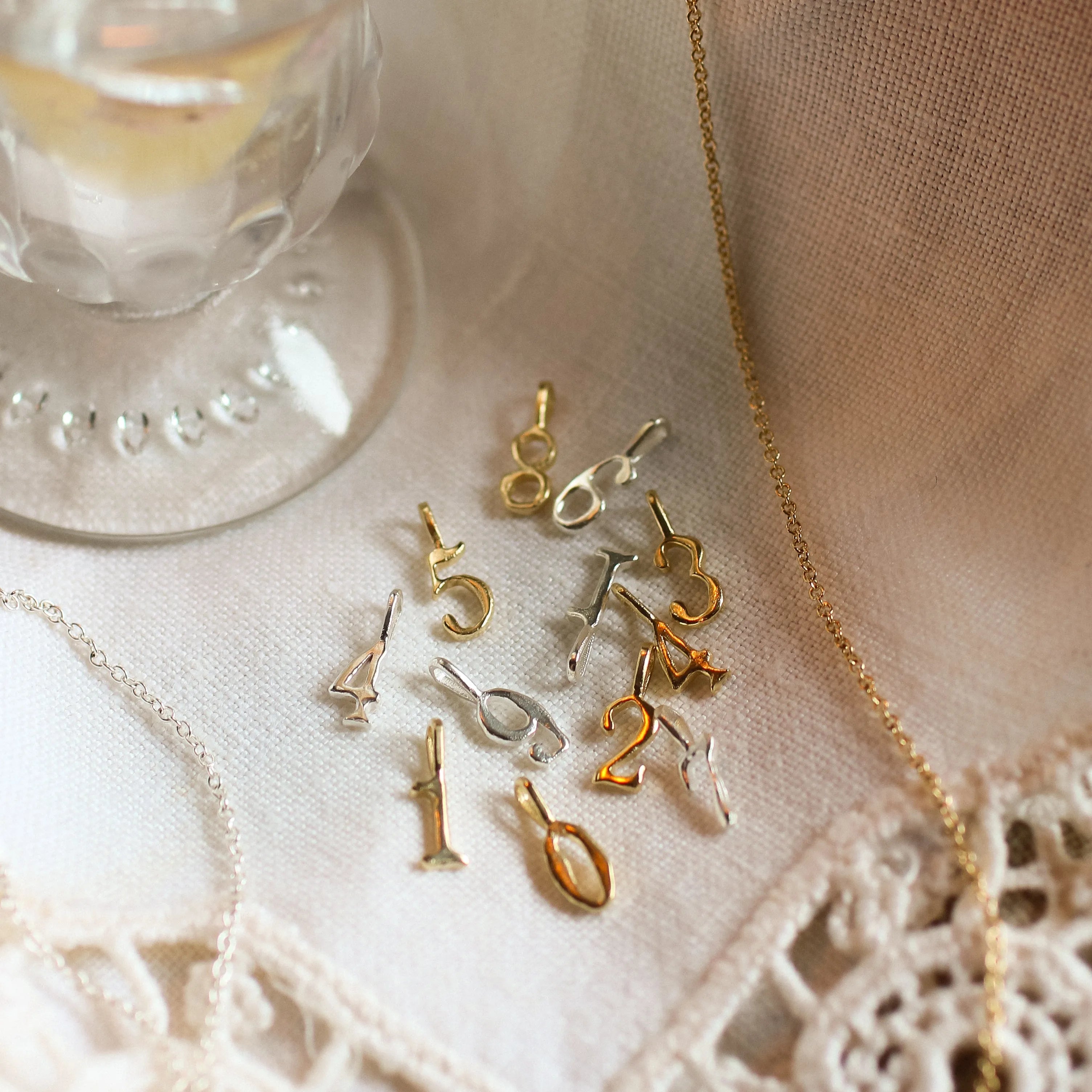 Gold jewelry with number charms on a lace tablecloth next to a candle