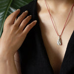 Person wearing a black top with a red necklace featuring a silver pendant, against a blurred green leaf background.