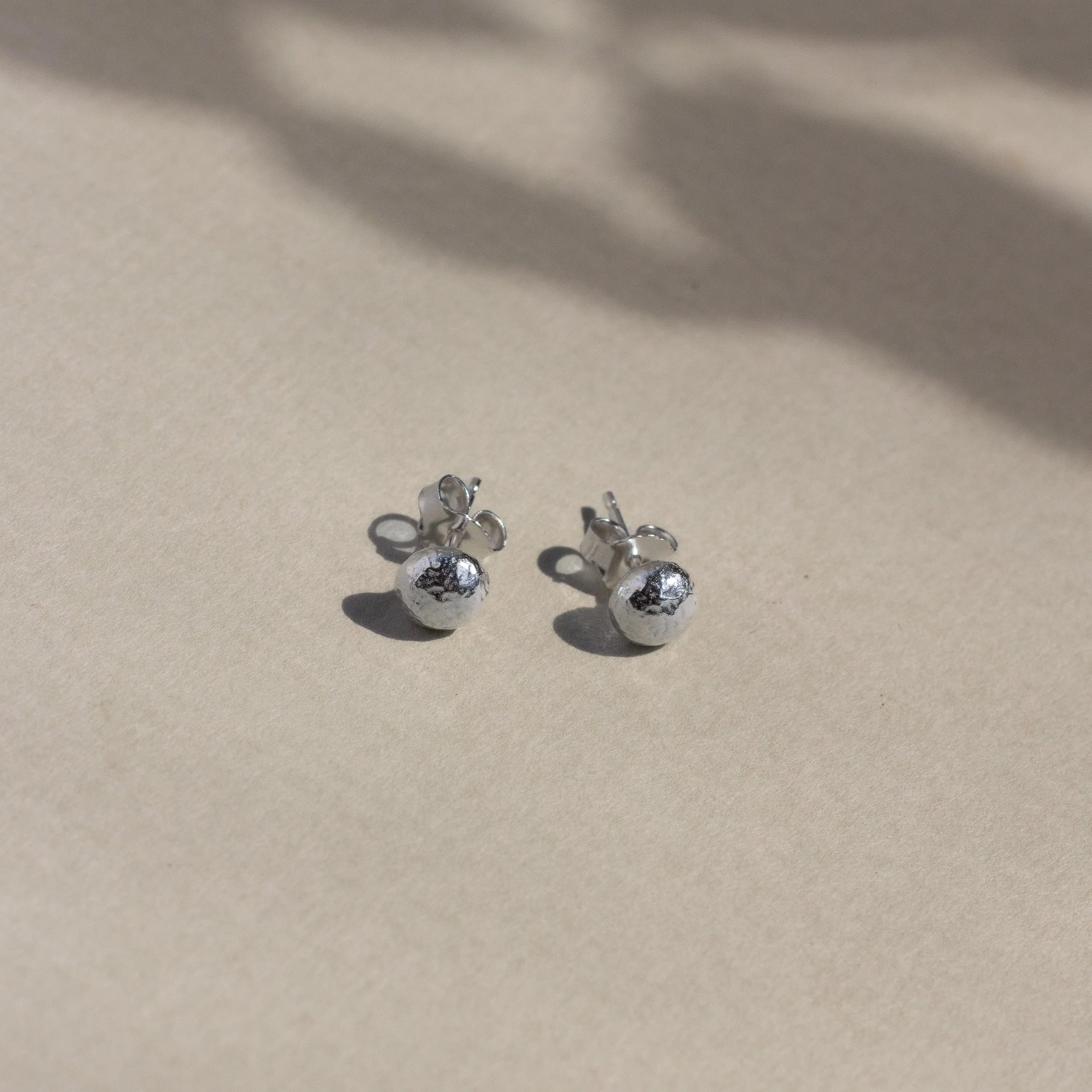 silver textured pebble-shaped earrings with butterfly backs on beige surface with leaf shadows