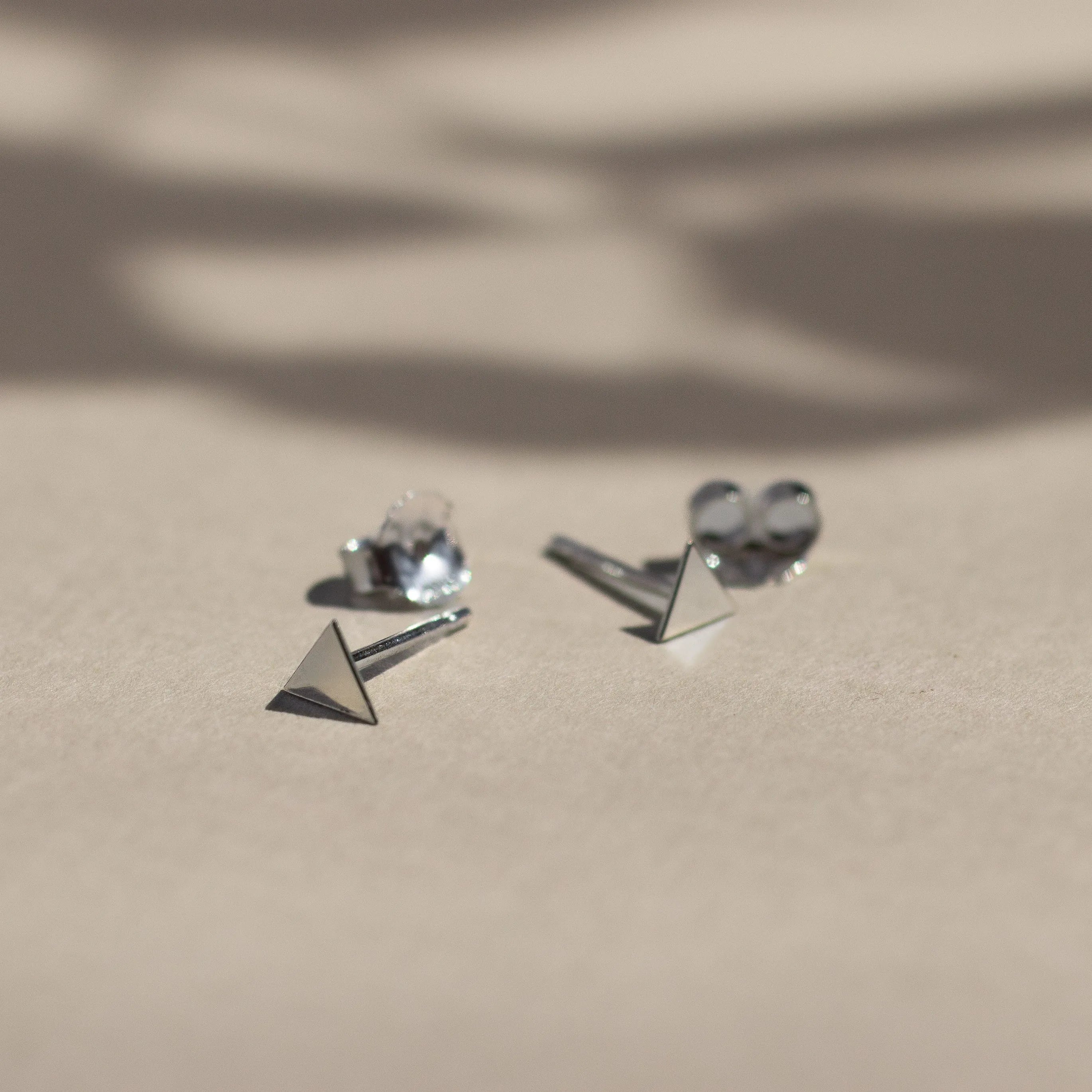 silver triangle stud earrings with butterfly backs on beige surface with leaf shadows
