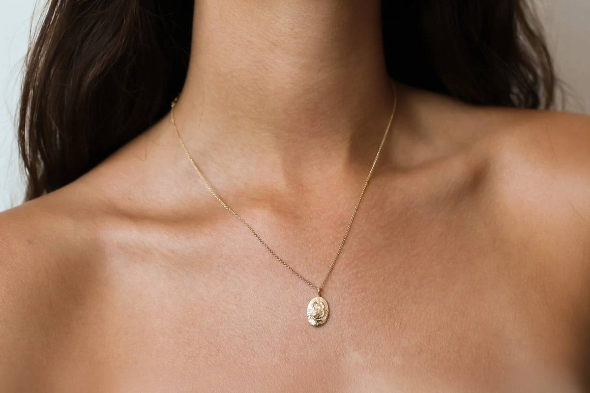 A close-up of a model's neck and upper chest wearing a delicate gold chain zodiac necklace with a small oval pendant featuring an embossed Cancer crab design.