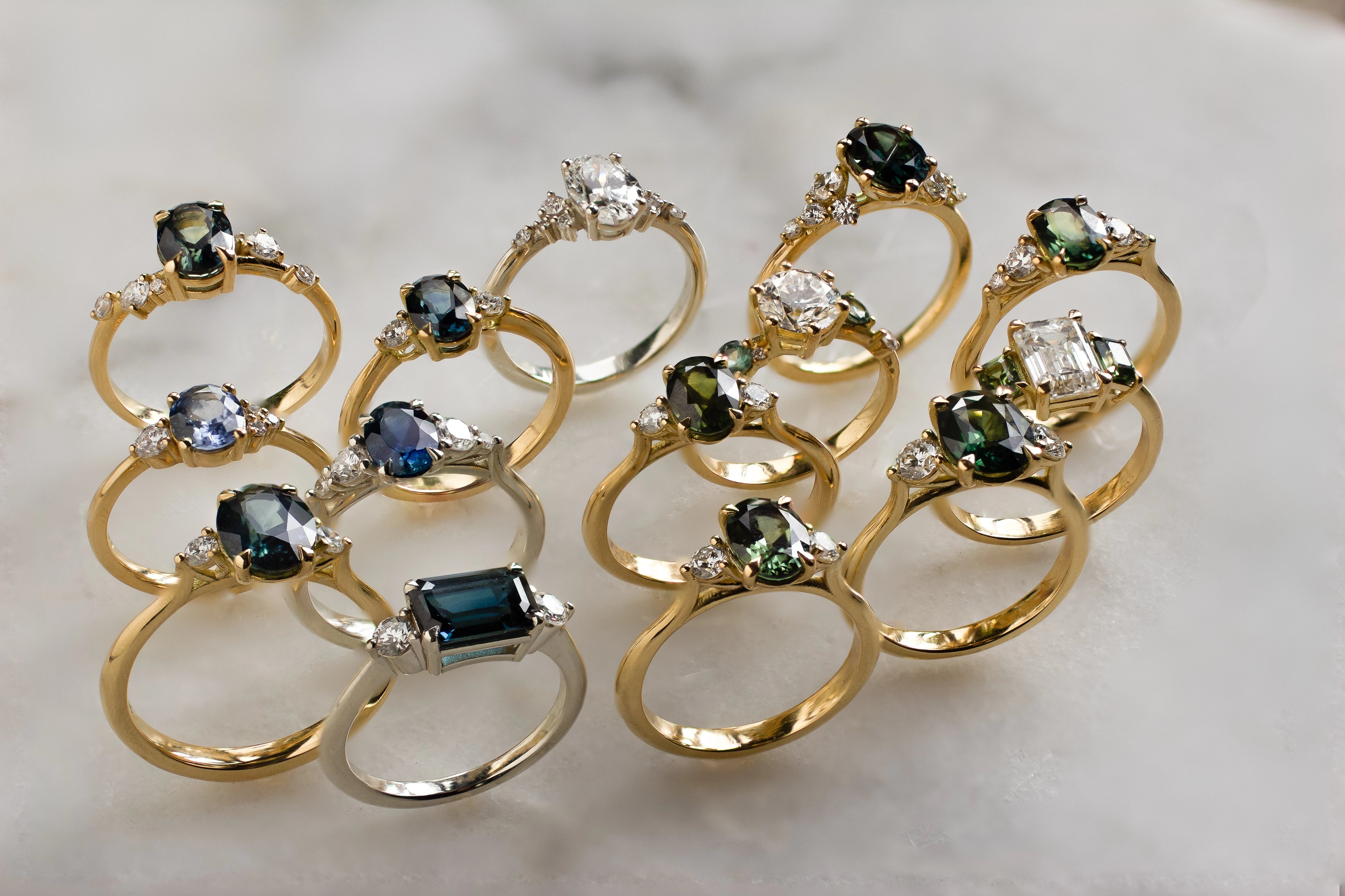 14 engagement rings with blue and green stones and diamonds on marble surface