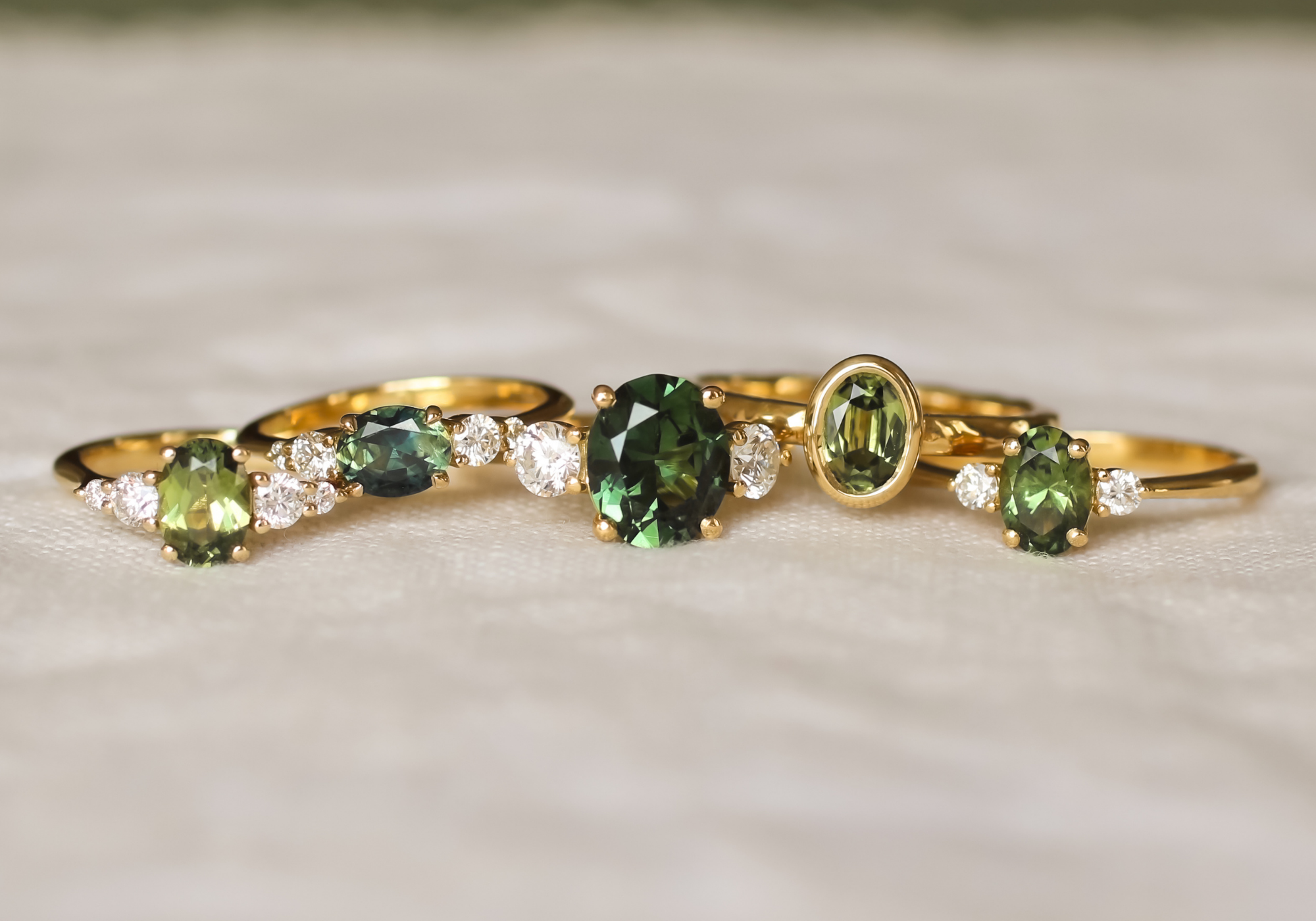 5 green sapphire engagement rings with diamonds on a beige surface and blurred background