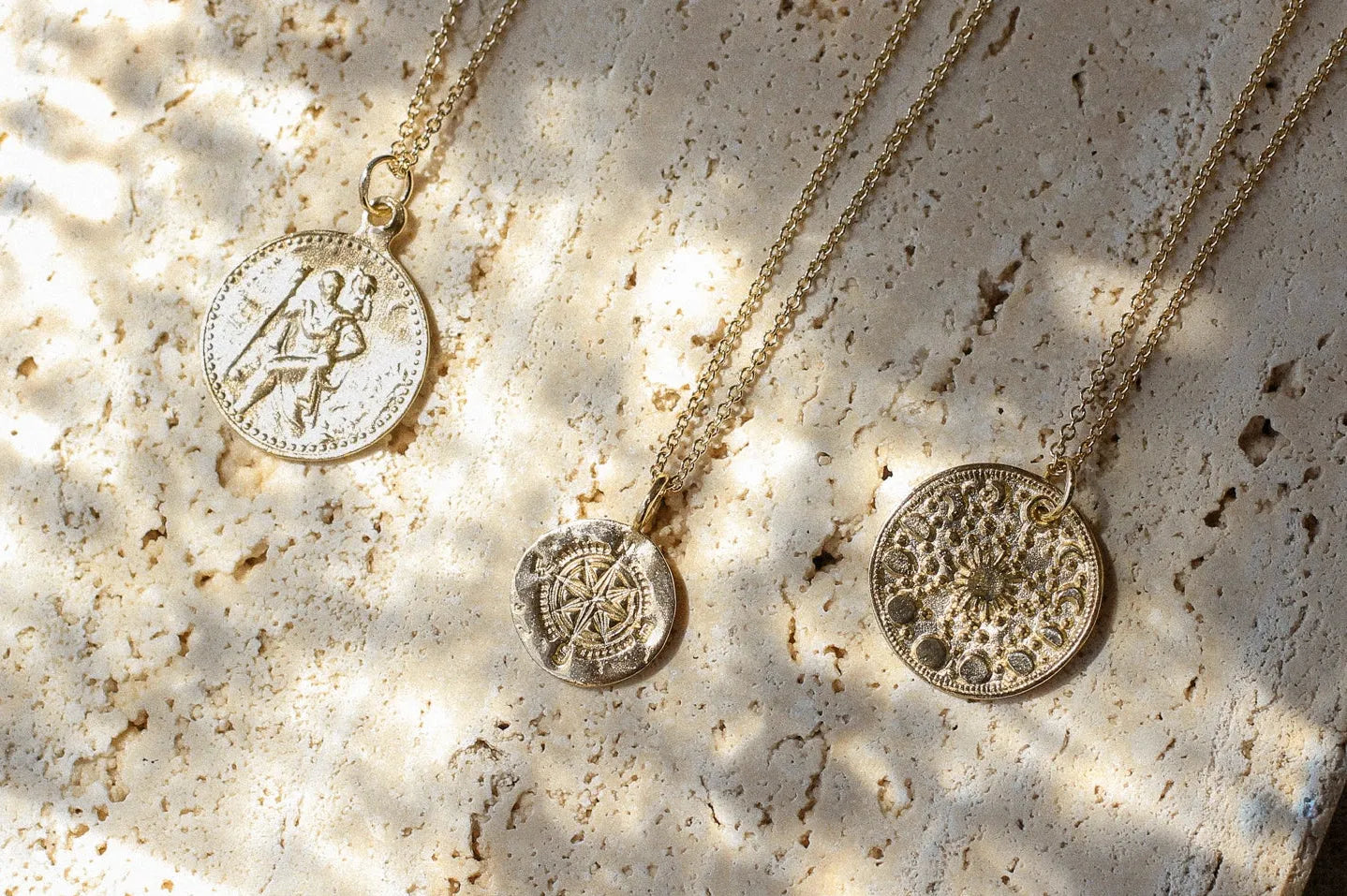 three gold necklaces with st christopher, compass and moonphase design on a stone block in the sun