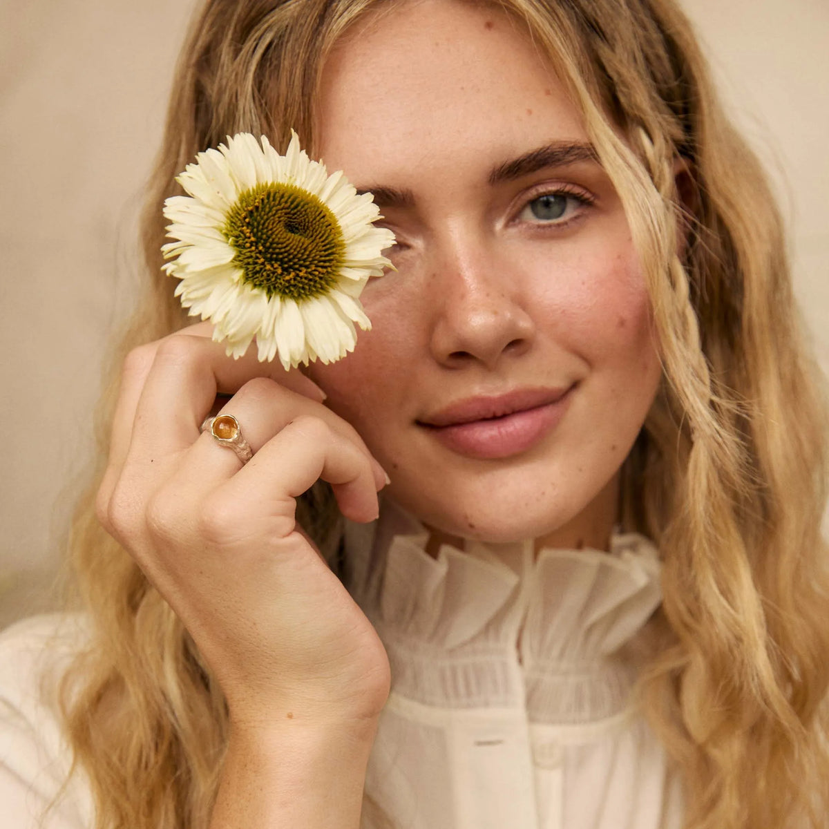 A woman with long blonde hair holds a white daisy flower in front of one eye, wearing a white ruffled blouse, a silver bracelet, and a gold ring with a citrine stone.