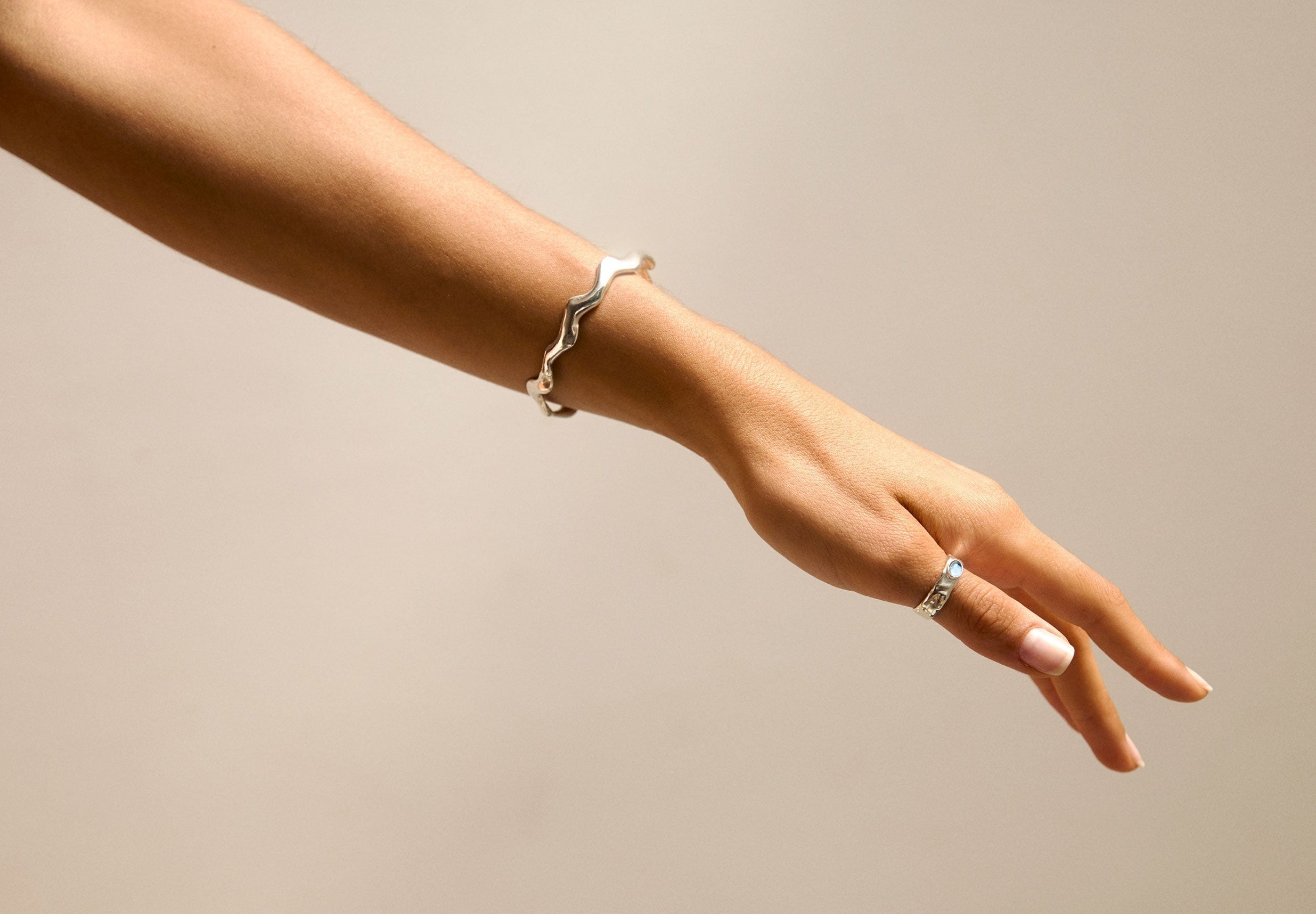 A person's hand and forearm wearing a silver wavy bangle on the wrist and a silver ring on the finger, set against a plain beige background.