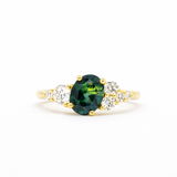 gold engagement ring with blue/green oval sapphire with asymmetric diamond cluster on white background