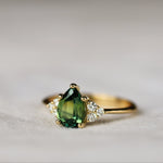 engagement ring with green pear-shaped sapphire and round diamonds on a fabric surface with blurred background