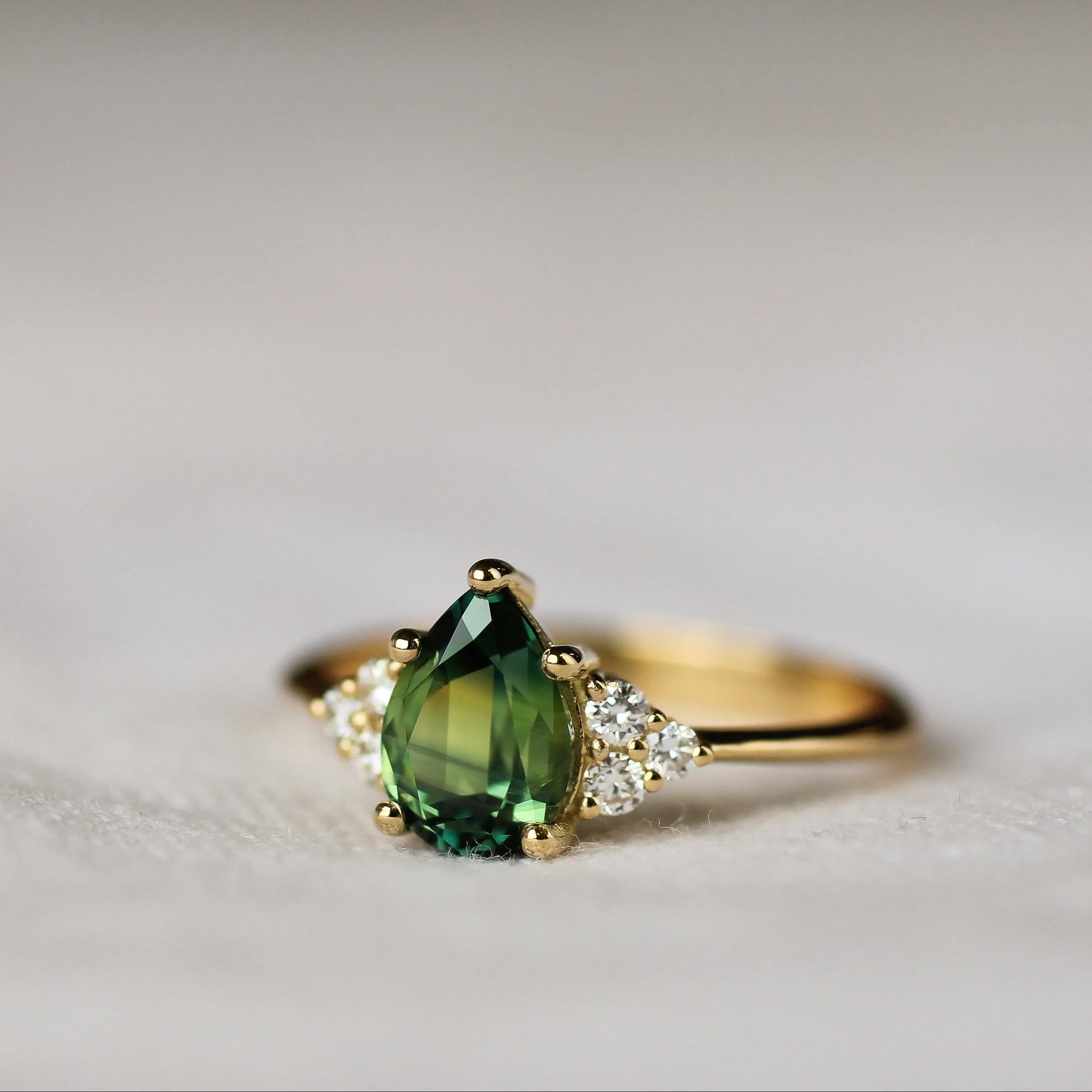 engagement ring with green pear-shaped sapphire and round diamonds on a fabric surface with blurred background