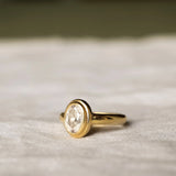 side view of double bezel oval diamond engagement ring with chunky band on a beige fabric and blurred background
