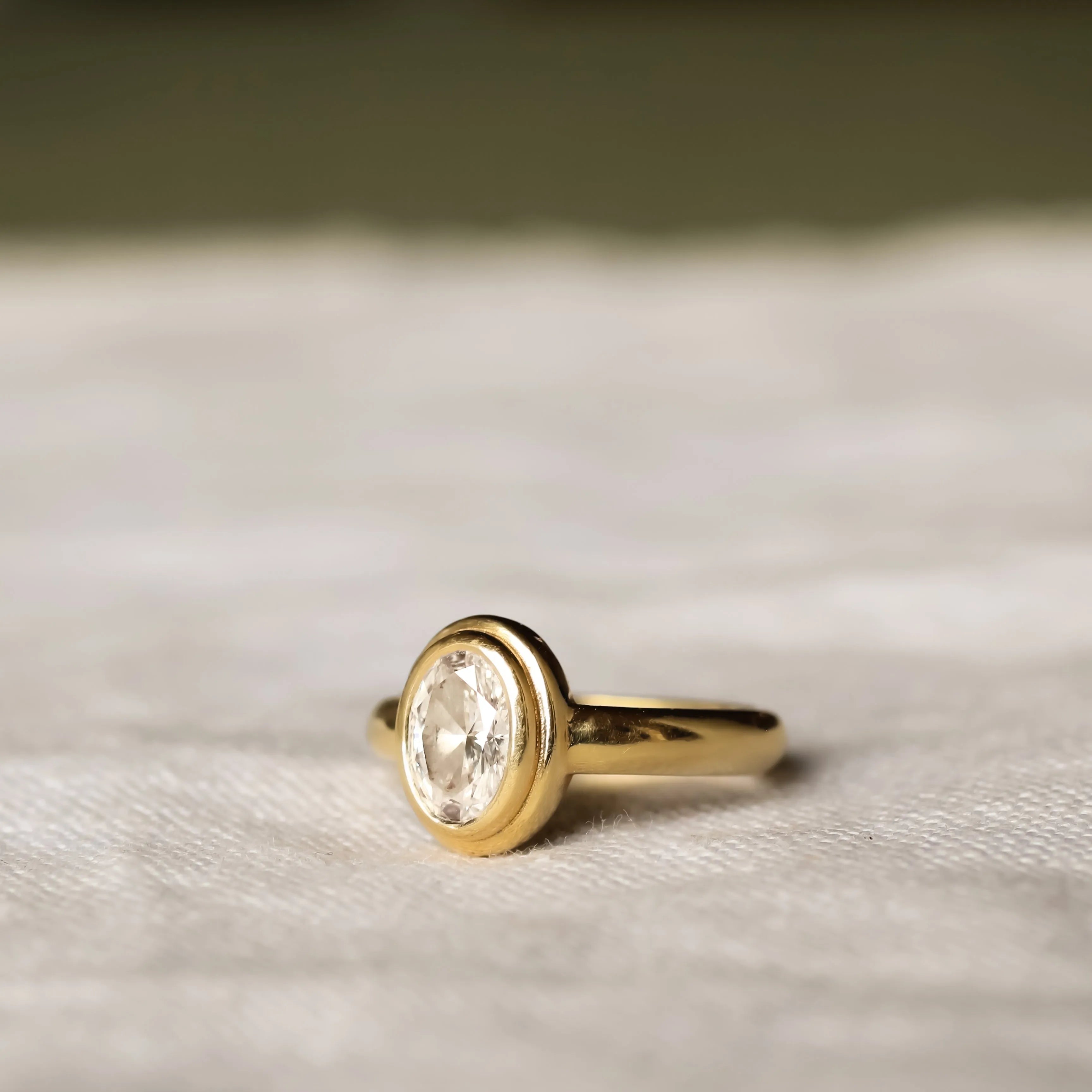 side view of double bezel oval diamond engagement ring with chunky band on a beige fabric and blurred background