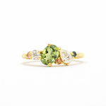 Gold ring with a green oval sapphire, pear diamond and pink and teal sapphires on a white background