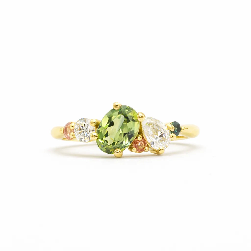 Gold ring with a green oval sapphire, pear diamond and pink and teal sapphires on a white background