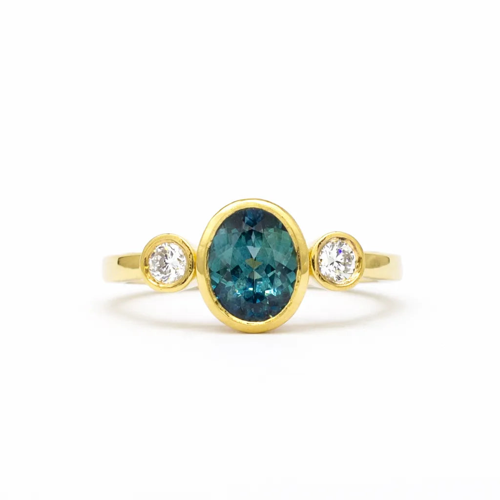 gold engagement ring with oval blue sapphire and round diamonds bezel set on a white background