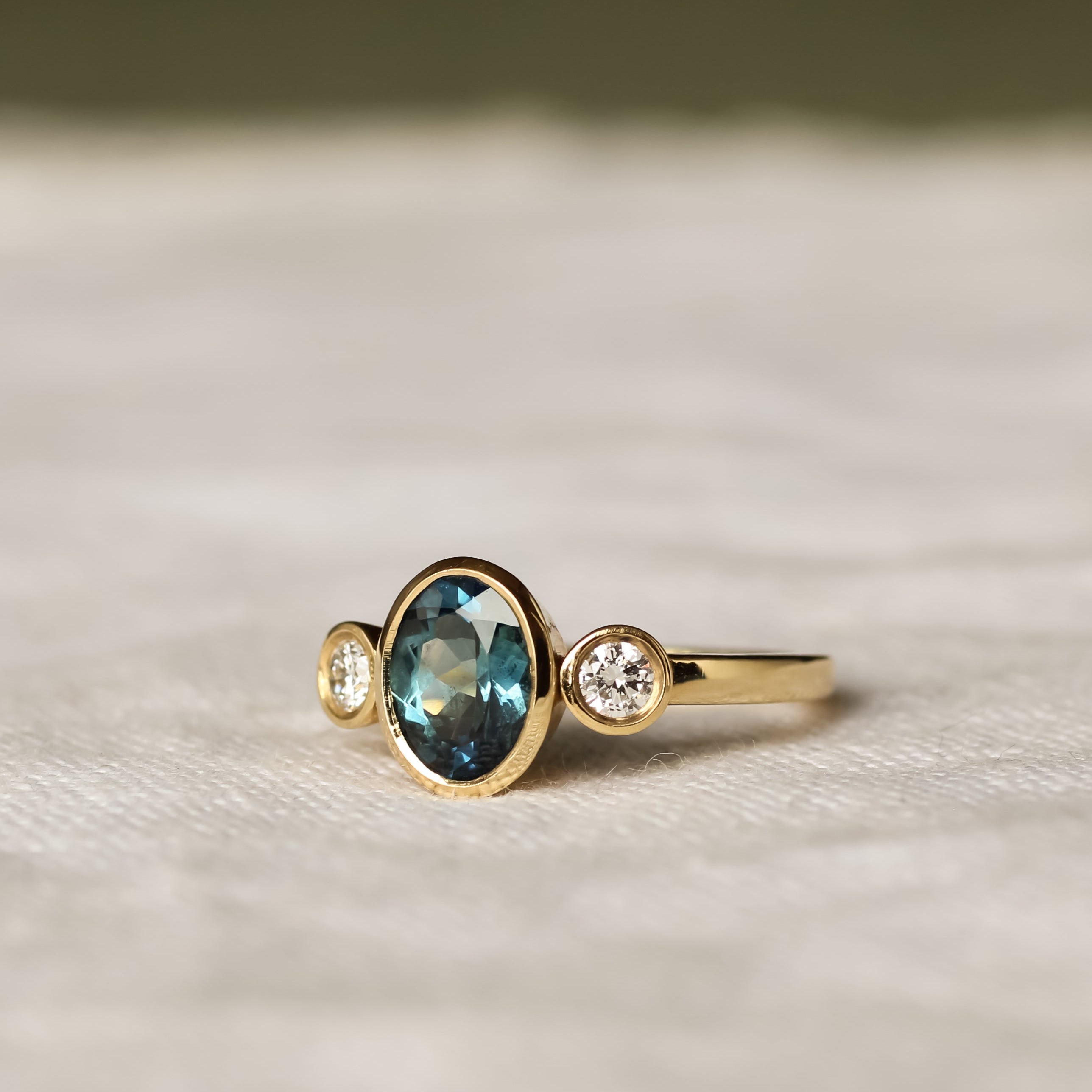 bezel set oval trilogy engagement ring with teal sapphire and round diamonds on a beige fabric