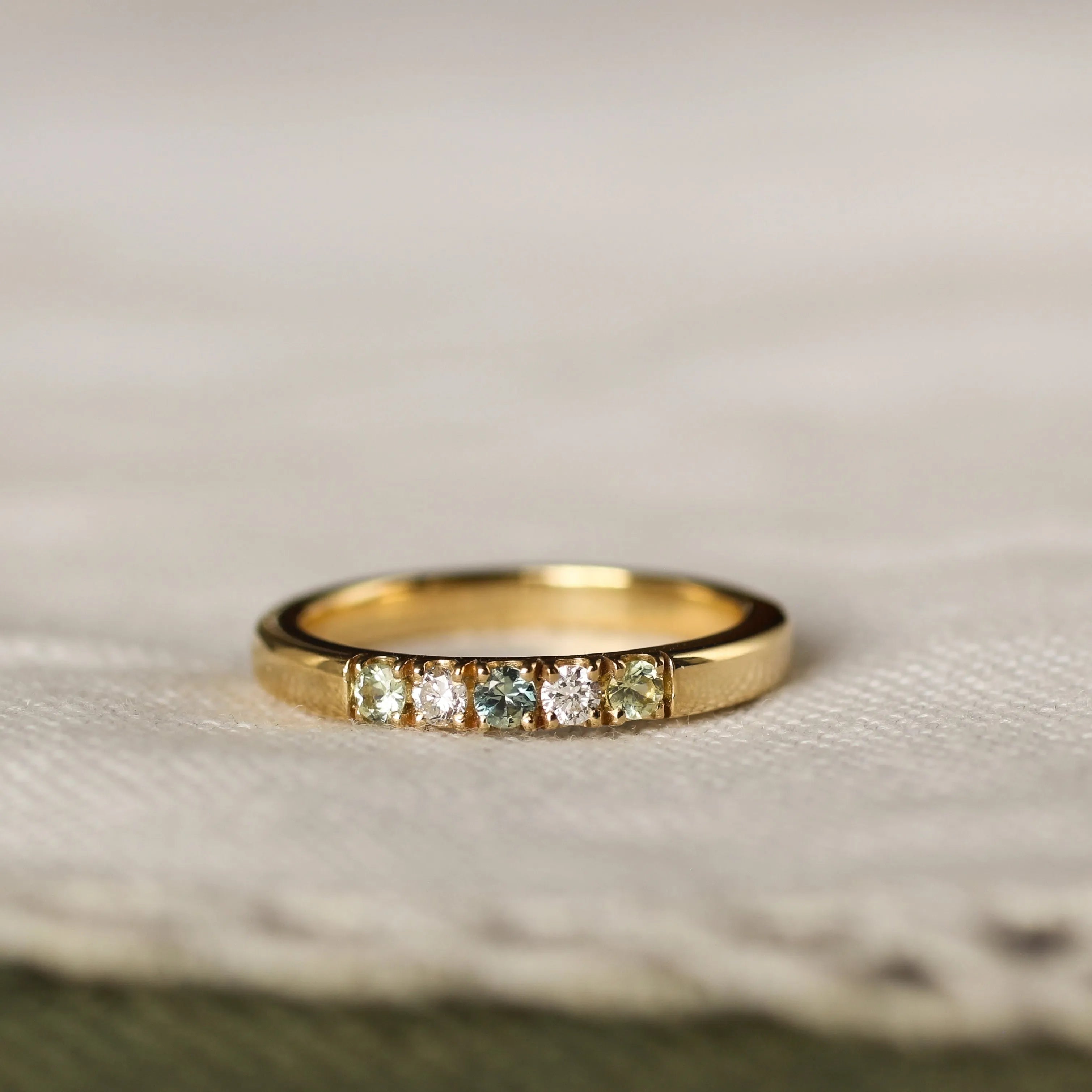 gold ring with 5 gemstones including diamonds and blue green sapphires on a beige fabric