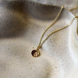 A delicate gold chain necklace with a small circular pendant resting on a textured beige ceramic dish.