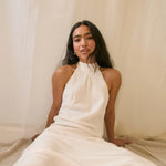 Woman wearing a sun dangle earrings and a white halter neck dress sitting against a beige curtain.